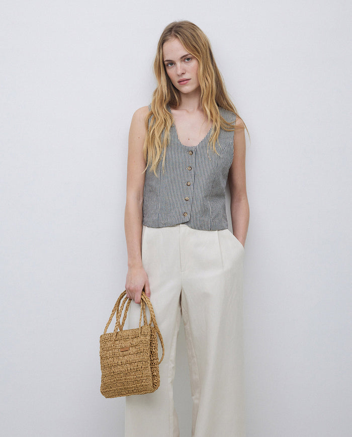 Woman wearing a sleeveless striped top and white pants, holding a straw bag against a plain background